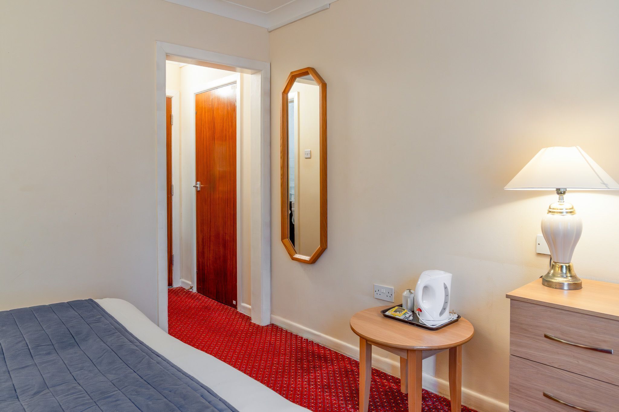 Una habitación de hotel con cama, espejo, lámpara, una mesita con tetera y piso alfombrado en rojo.