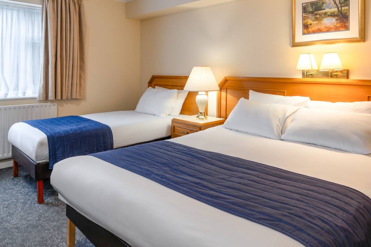A hotel room with two beds, a nightstand with a lamp, a window with curtains, and a painting on the wall.