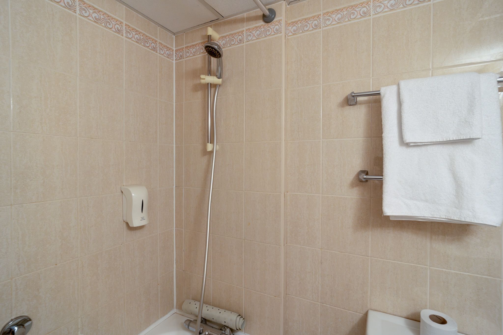 A bathroom with beige tiles, a showerhead, a towel rack with two towels, and a toilet paper holder.