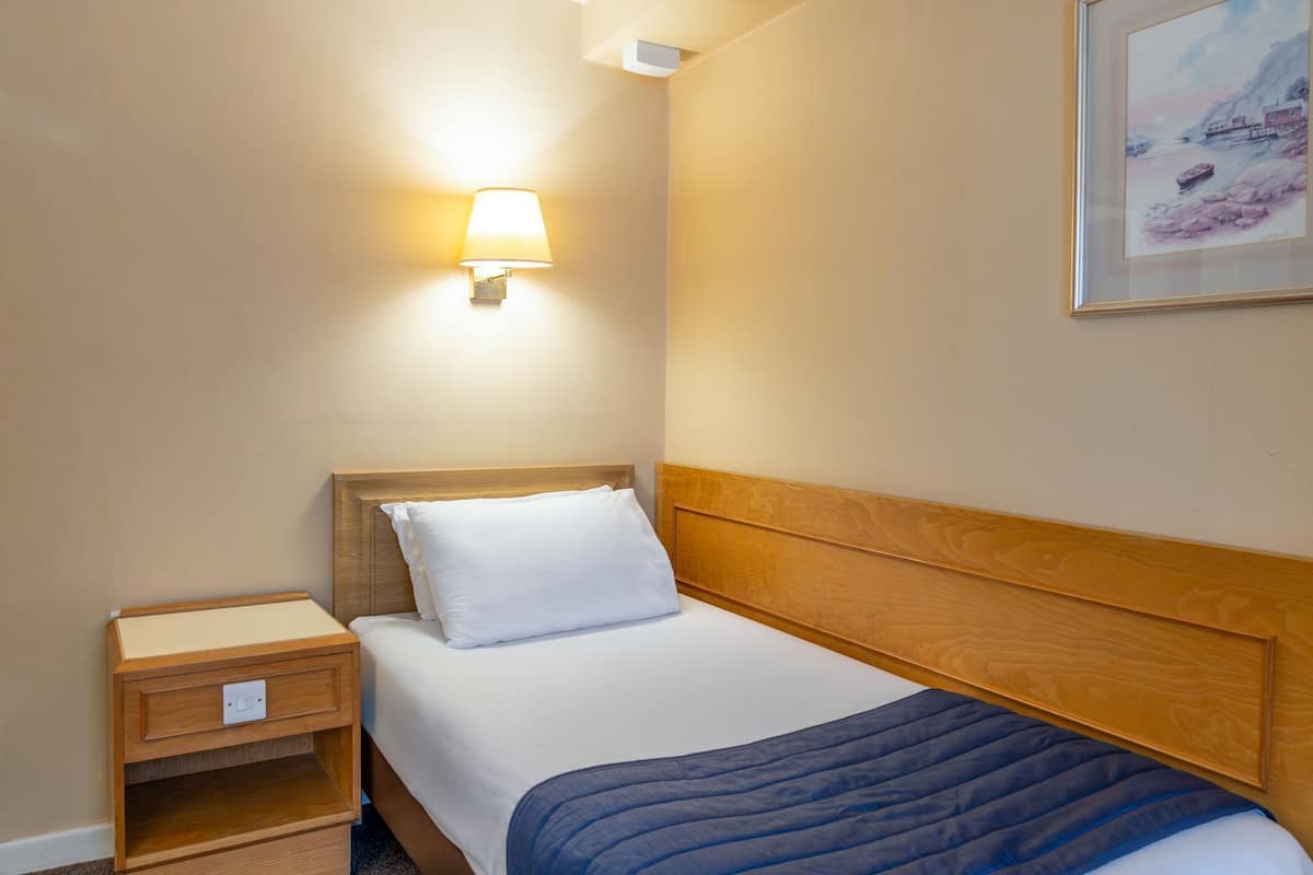A hotel room with a bed, a nightstand, a wall-mounted lamp, and a framed picture on the wall.