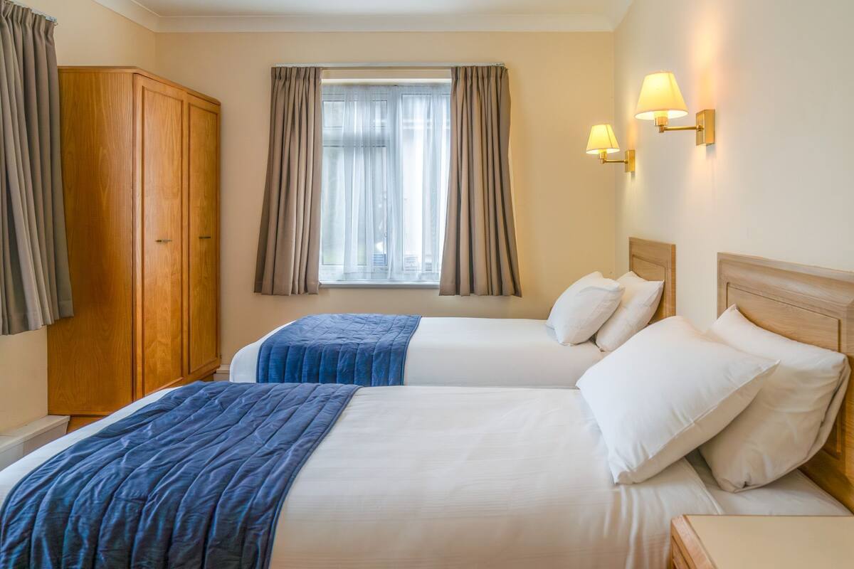 A hotel room with two beds, a wardrobe, a window with curtains, and wall-mounted lamps.