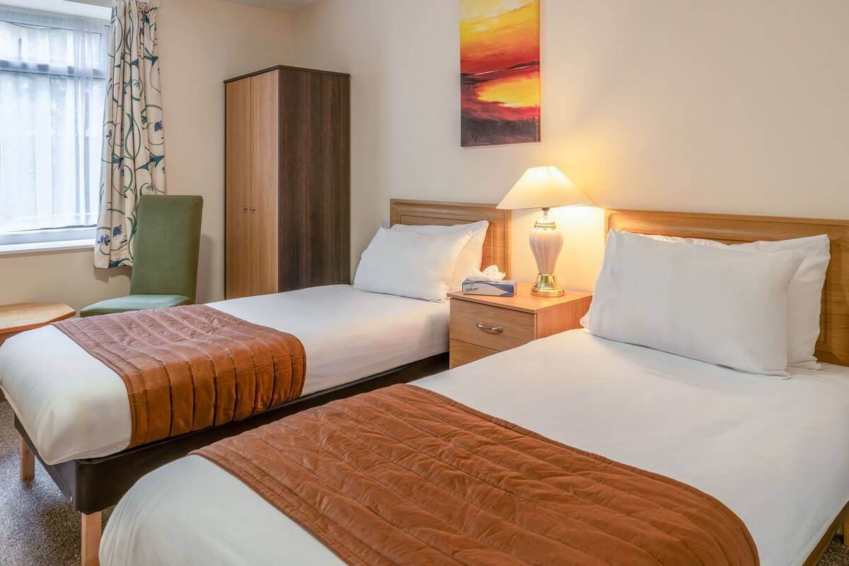A hotel room with two beds, a wooden wardrobe, a green chair, a window with curtains, and a sunset painting on the wall.