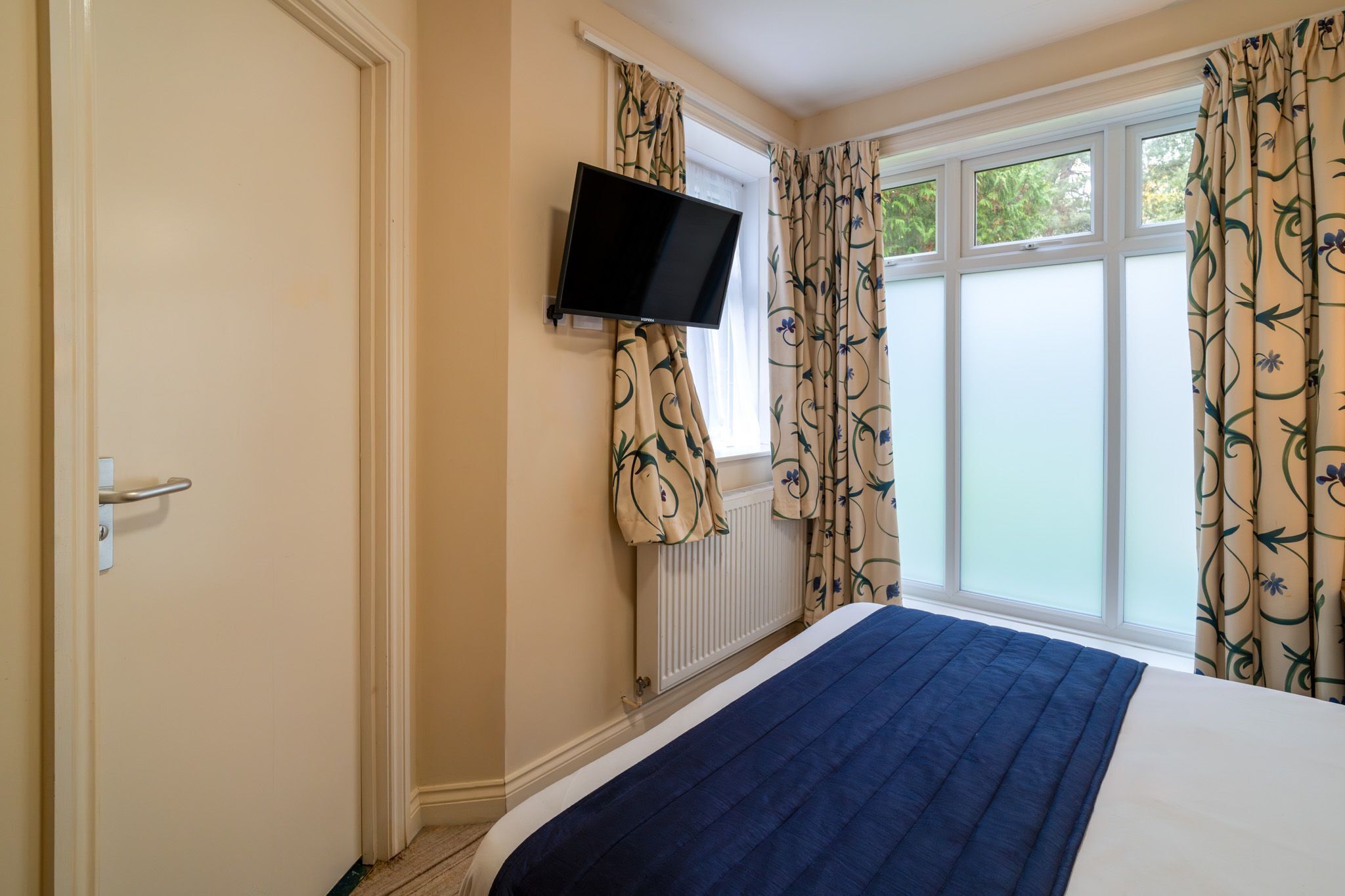 Une chambre à coucher avec un lit, un téléviseur fixé au mur et une fenêtre avec des rideaux.
