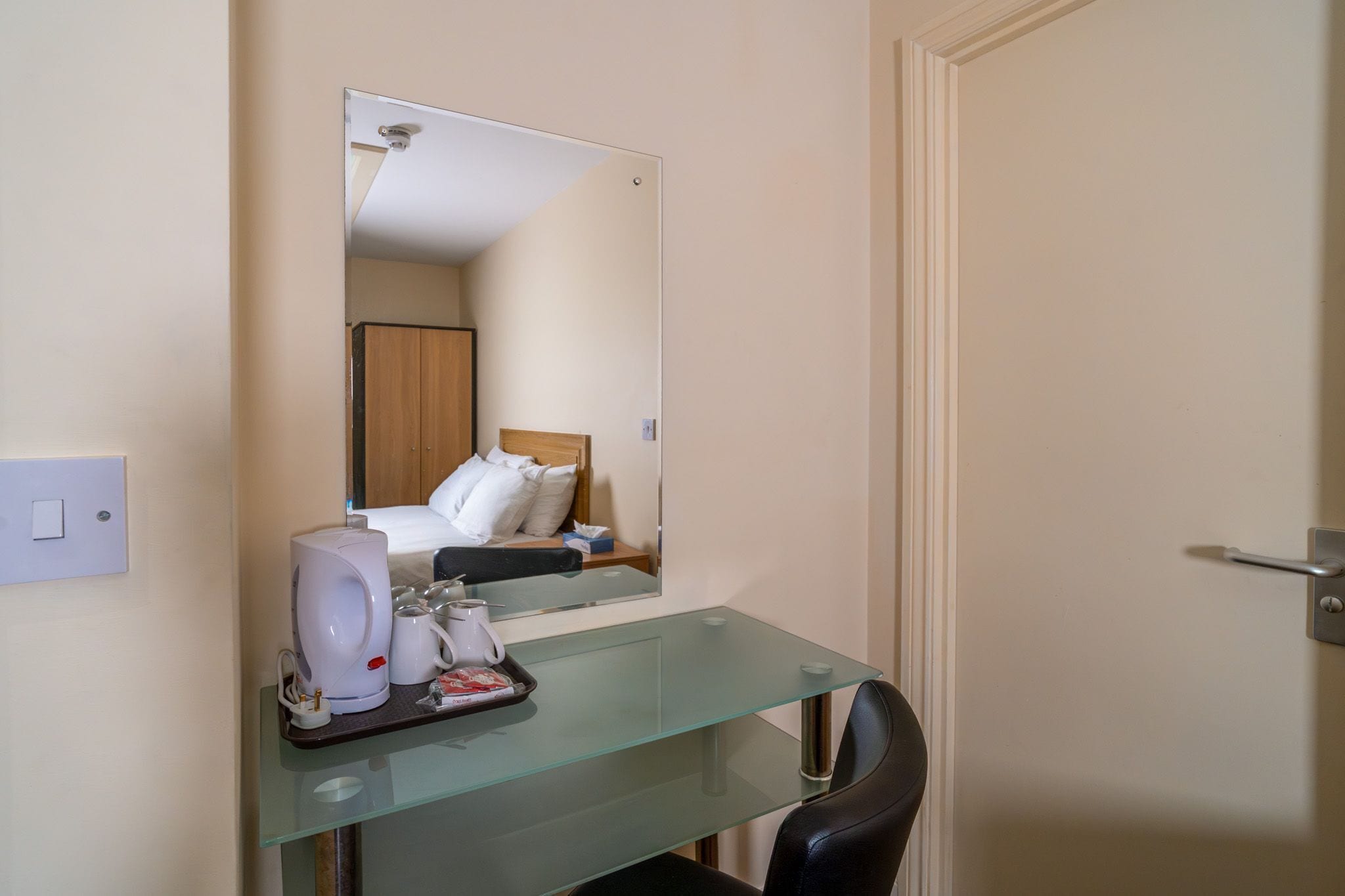 Quarto de hotel com escrivaninha de vidro, cama com roupa de cama branca, espelho, cafeteira e bandeja com objetos.