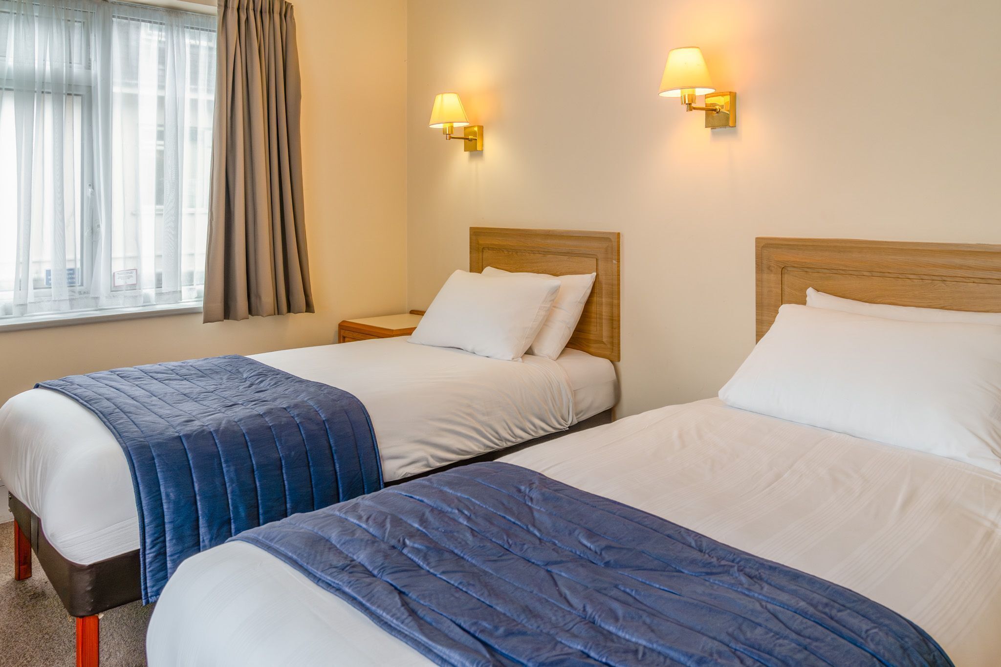 A hotel room with two beds, a window with curtains, and wall-mounted lamps.