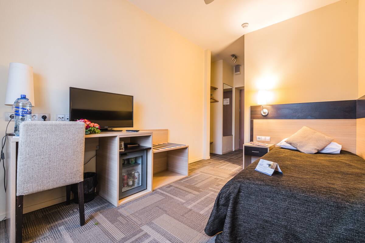 Quarto de hotel com cama, escrivaninha com televisão, armário pequeno e mesa de cabeceira com abajur.