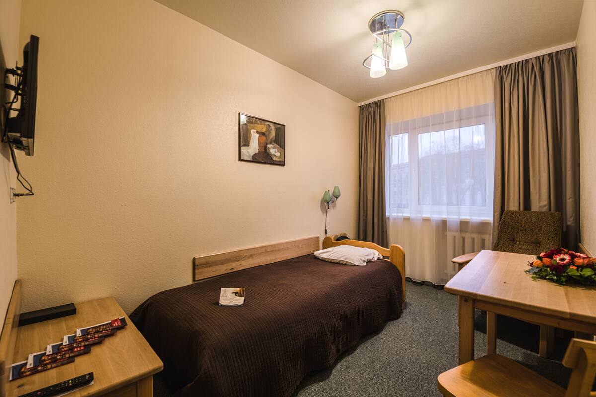 Quarto de hotel com uma cama de solteiro, uma mesa de cabeceira de madeira com um controle remoto, uma pequena mesa com um arranjo de flores e uma janela com cortinas.