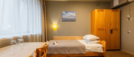A hotel room with a wooden bed, a dining table with chairs, a wall-mounted lamp, and a painting.