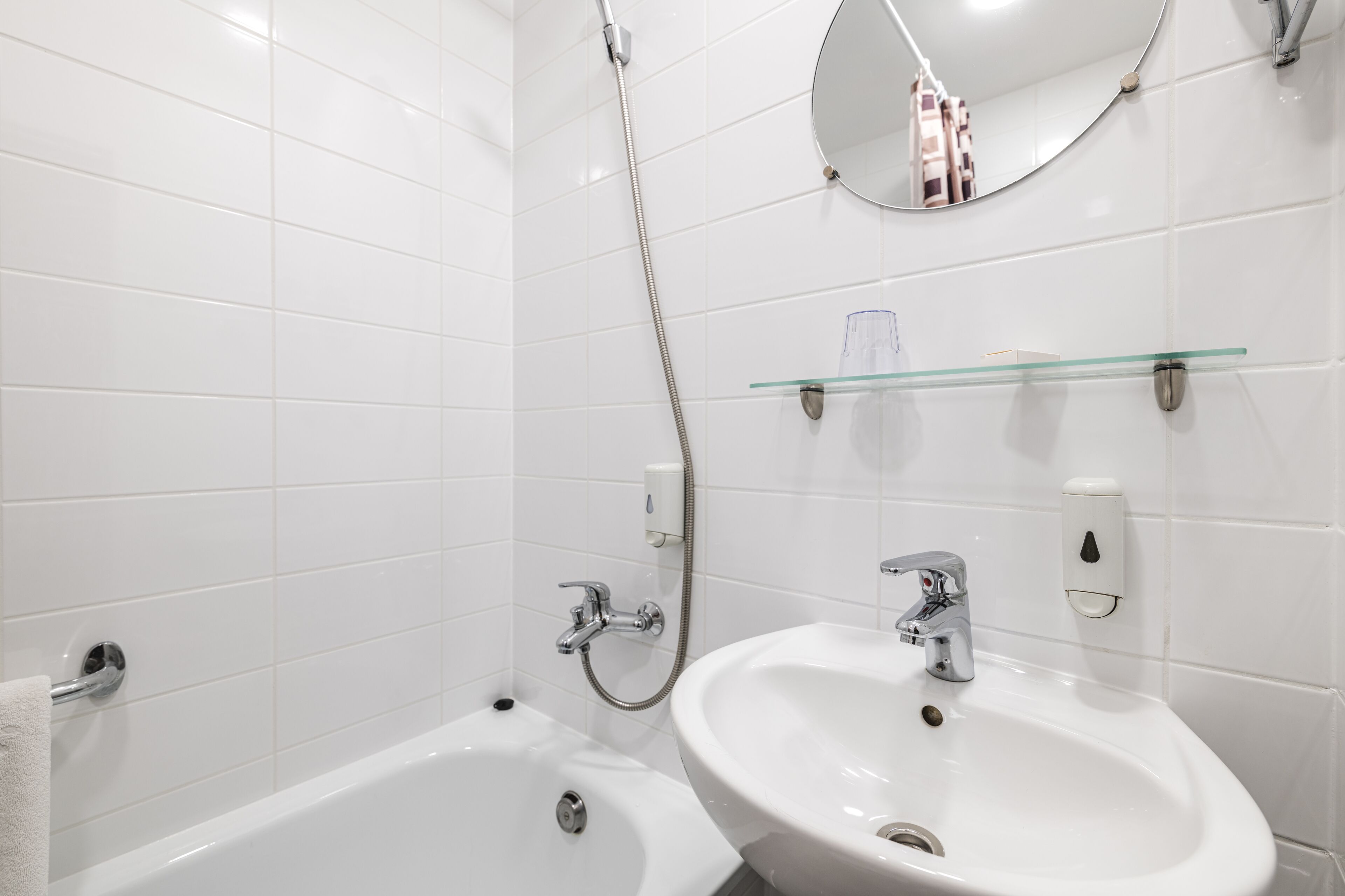 Un baño moderno con azulejos blancos, una bañera, un lavabo, una estantería de cristal y un espejo.
