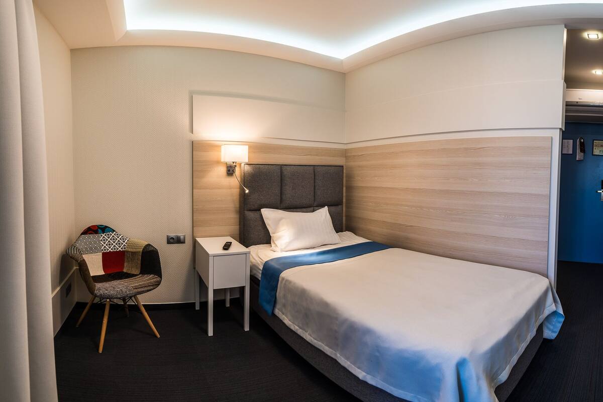 Una habitación de hotel con cama, mesita de noche, sillón y lámpara de pared.