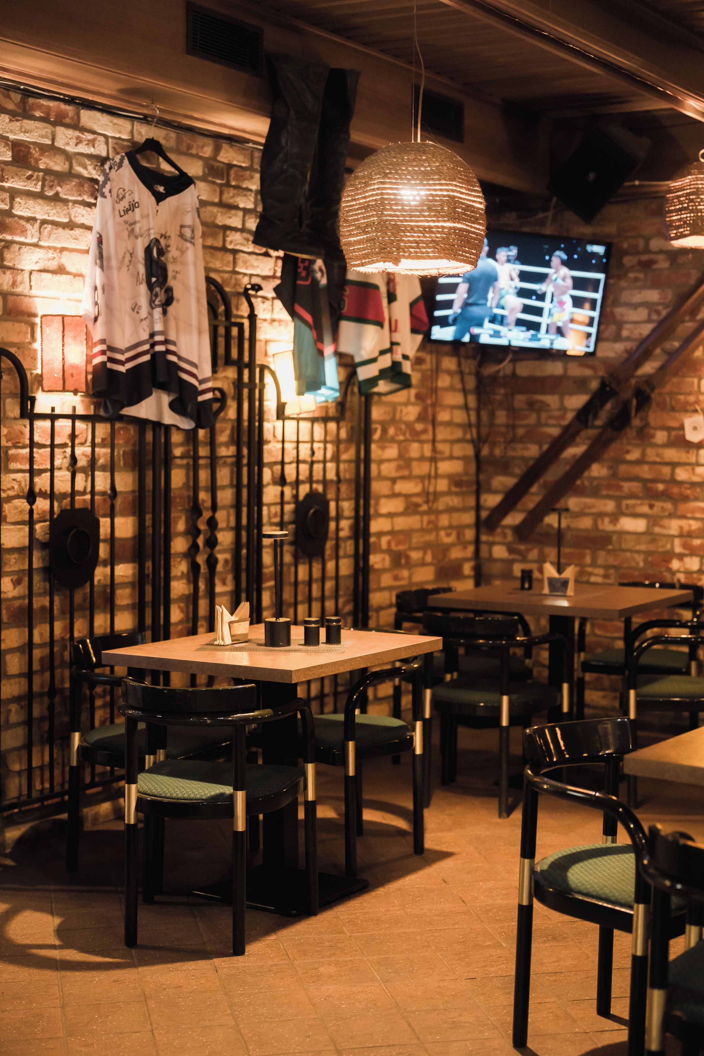 Interior de restaurante con paredes de ladrillo, camisetas colgadas, una pantalla de televisión y mesas y sillas de madera.