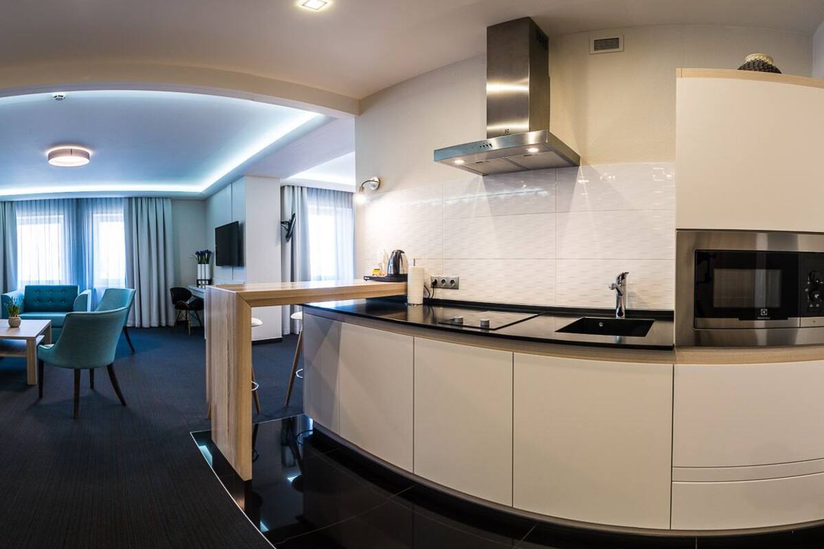 Una habitación de hotel moderna con una cocina curva, una zona de comedor con una mesa redonda y sillas, un sofá y un televisor.