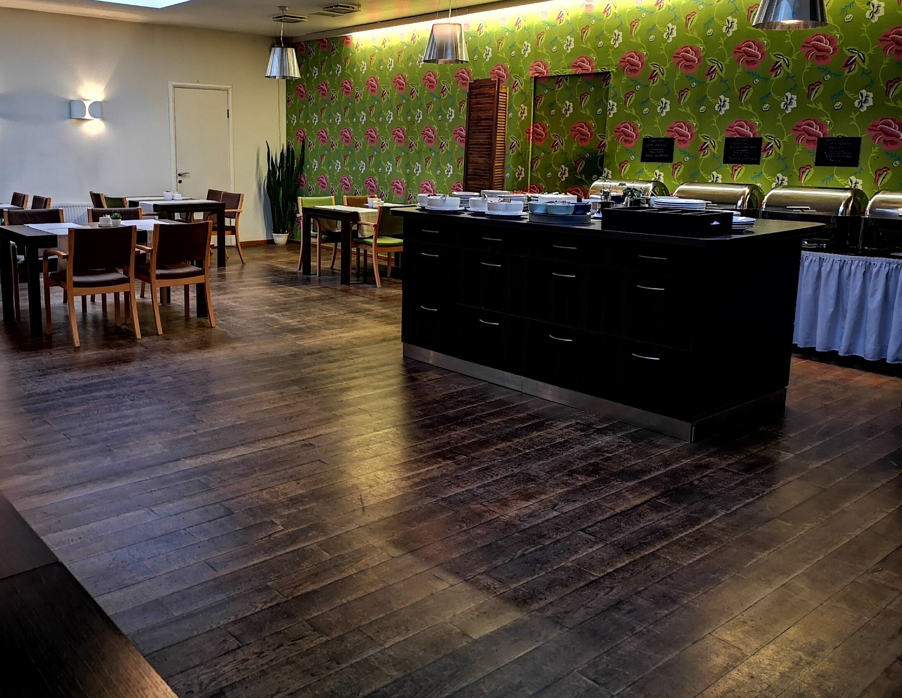 Un restaurante con suelo de madera, una isla central con encimera negra y un papel tapiz floral verde. Hay mesas y sillas dispuestas alrededor de la isla.