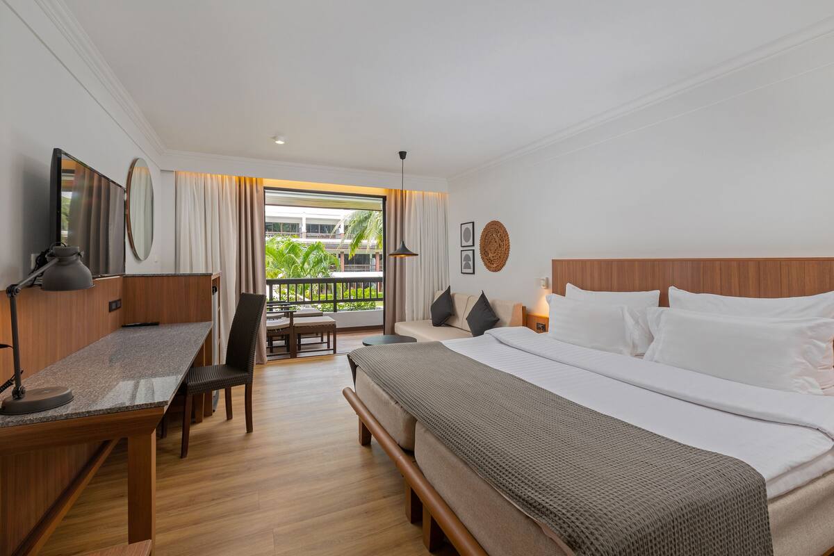 A hotel room with a bed, desk, chair, and a balcony with a view of greenery.