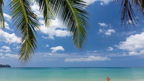 A beach with clear turquoise water, a sandy shore, and palm tree fronds framing the view. - Sunprime Kamala Beach (Kamala)