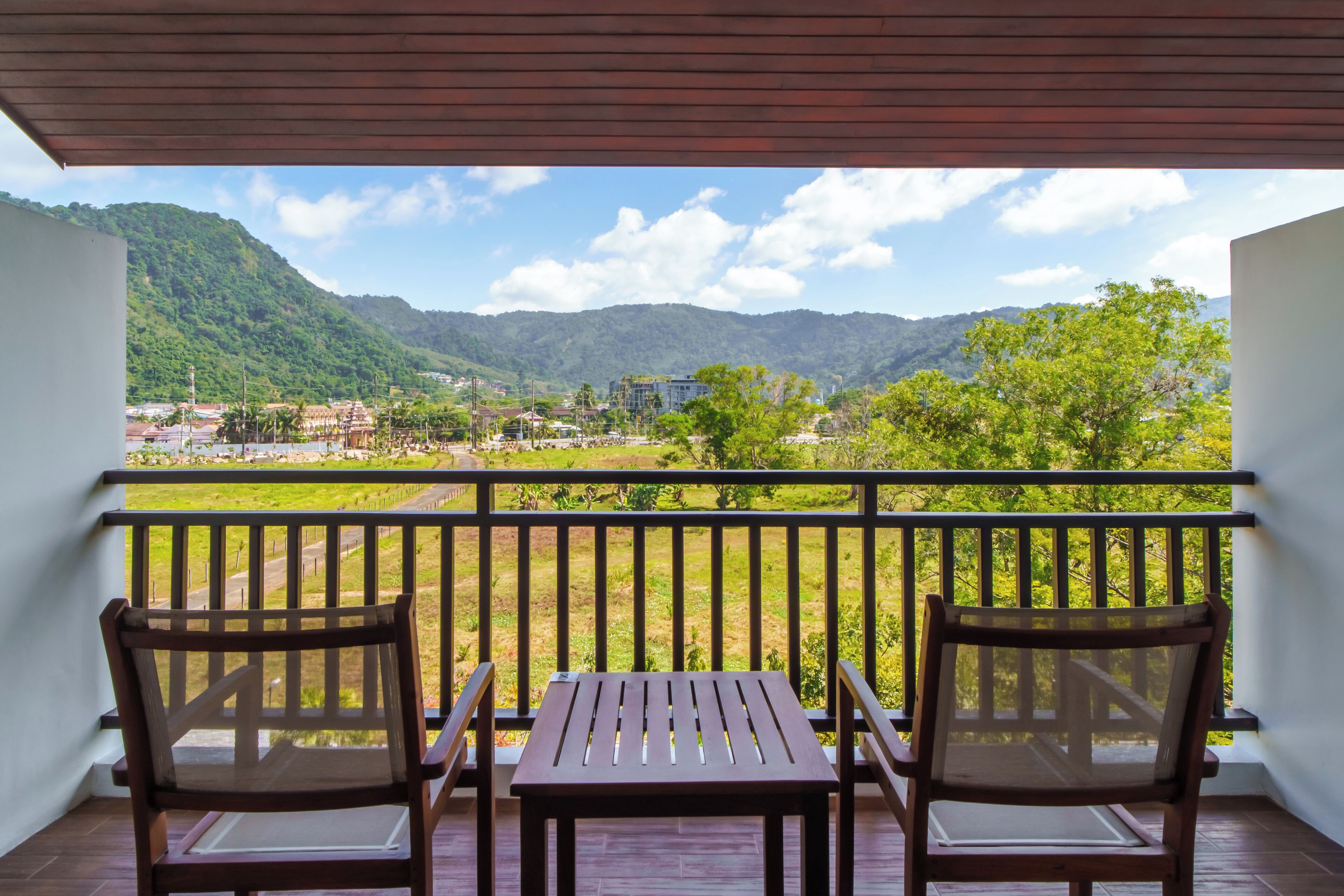 A balcony with two wooden chairs and a table, overlooking a green landscape with hills and buildings.