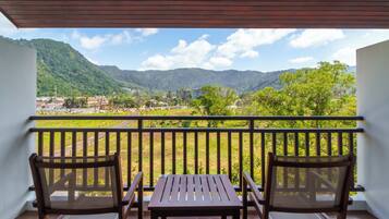 A balcony with two wooden chairs and a table, overlooking a green landscape with hills and buildings.