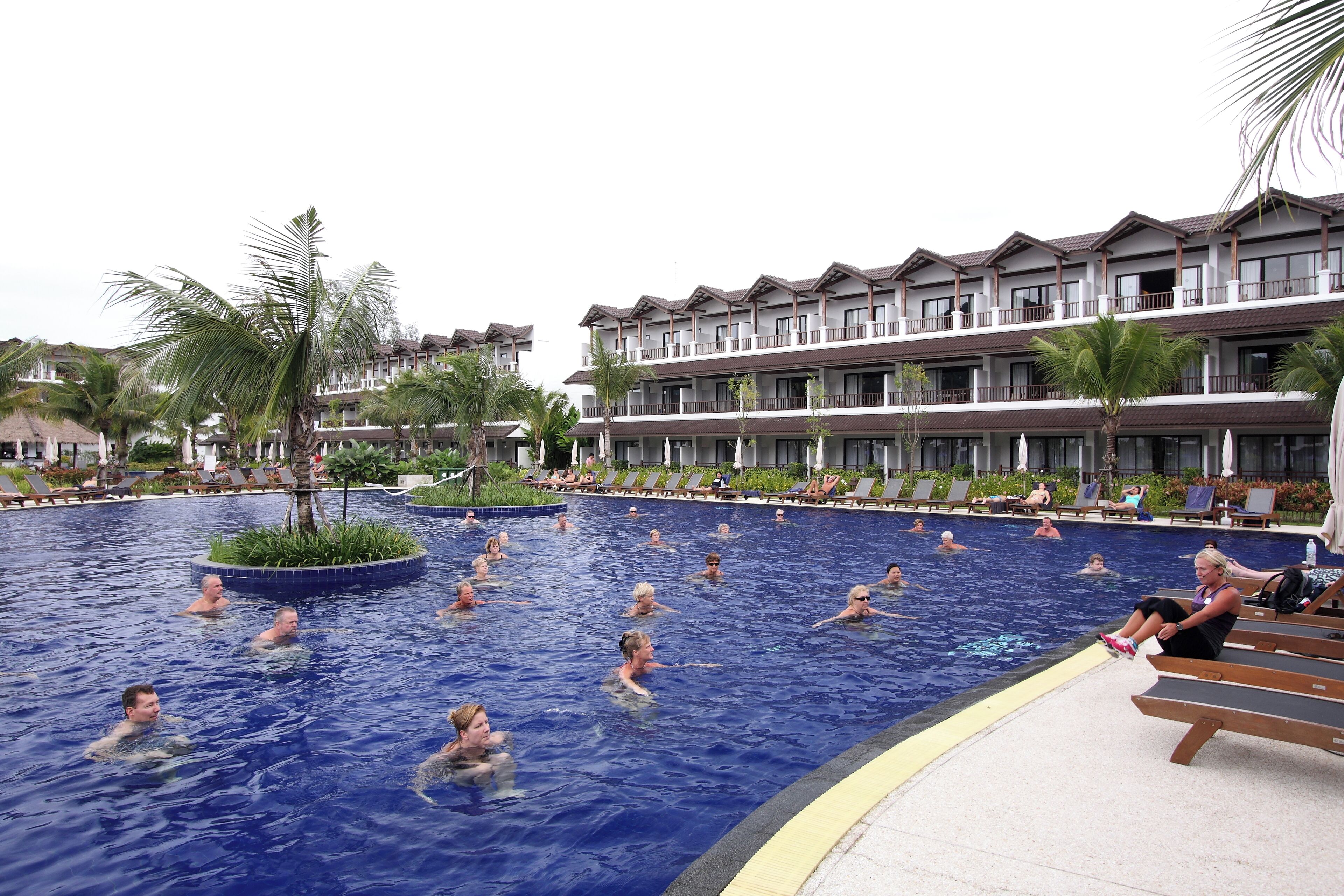 A swimming pool with people in the water, surrounded by lounge chairs and palm trees.