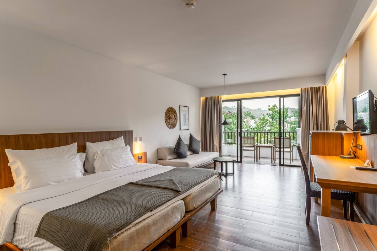 A hotel room with a large bed, a desk, a chair, a sofa, and a balcony with a view.