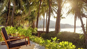 A wooden deck with a lounge chair and a woven bag, surrounded by lush greenery and palm trees, overlooking a beach. - Sunprime Kamala Beach (Kamala)
