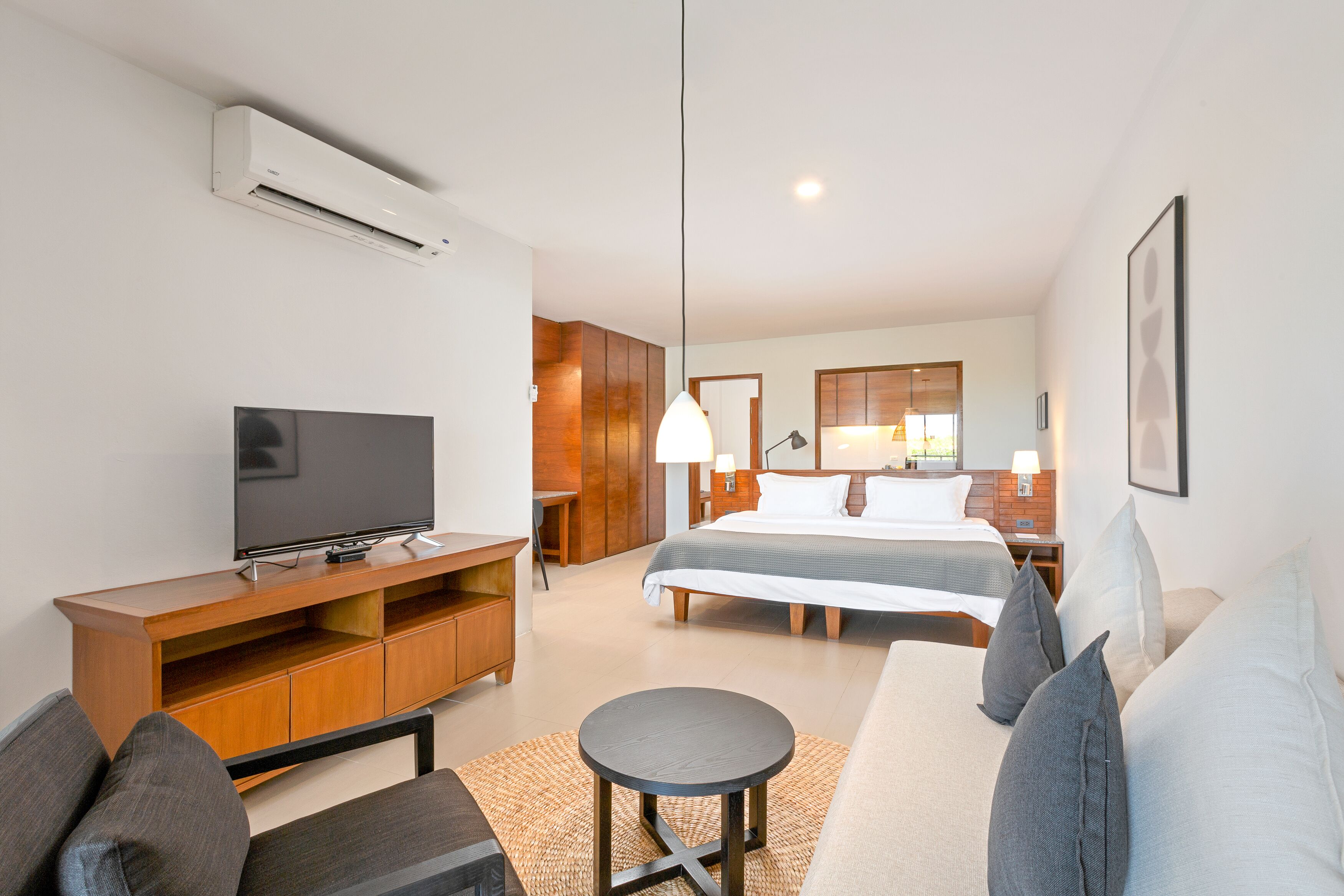 A modern hotel room with a flat-screen TV, a sofa, a round coffee table, a bed with two pillows, a nightstand with a lamp, and a wooden cabinet.