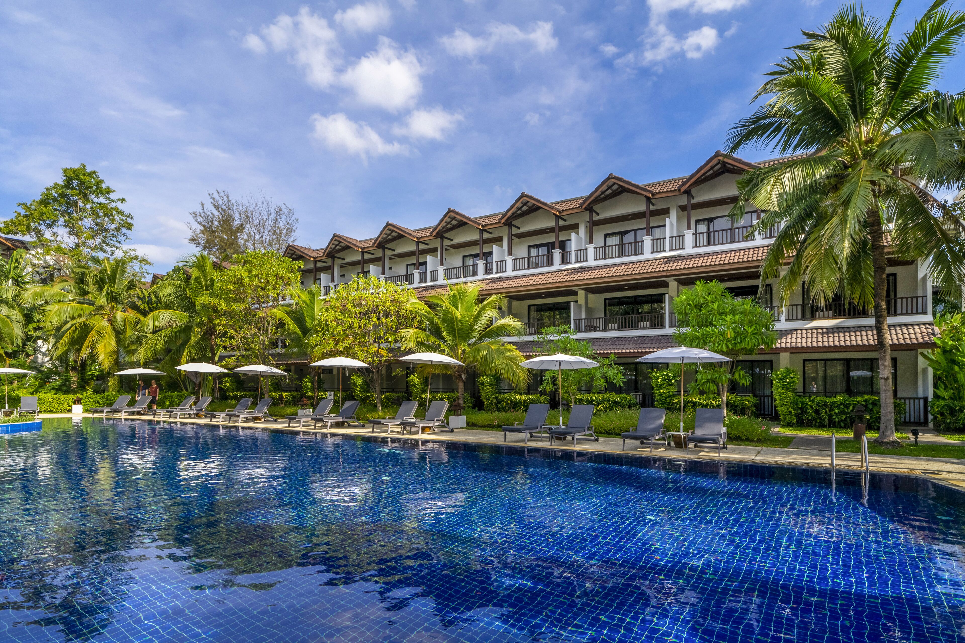 A hotel with a swimming pool, lounge chairs, umbrellas, and palm trees.