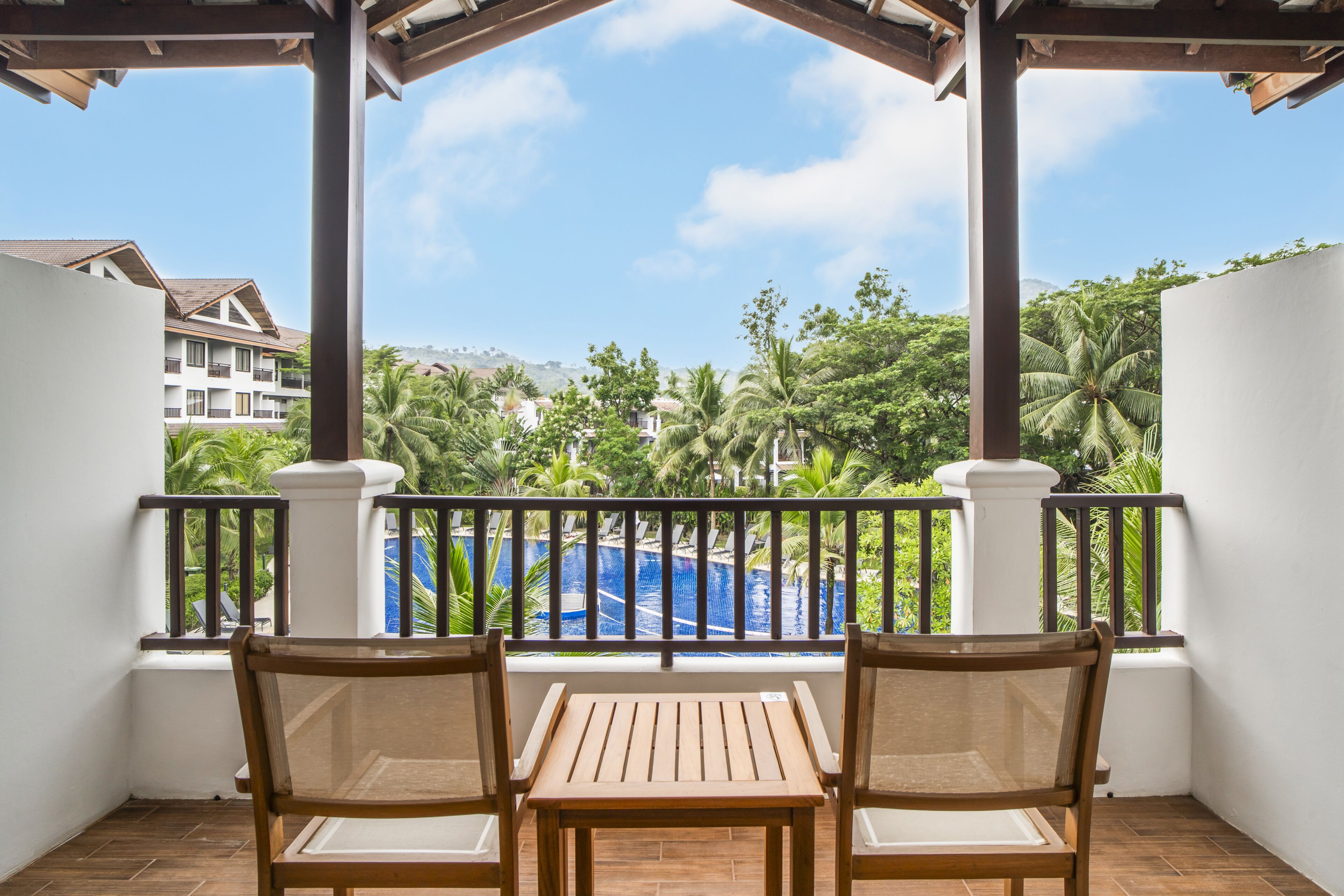 Un balcon avec deux chaises en bois et une table, donnant sur une piscine et une végétation luxuriante.