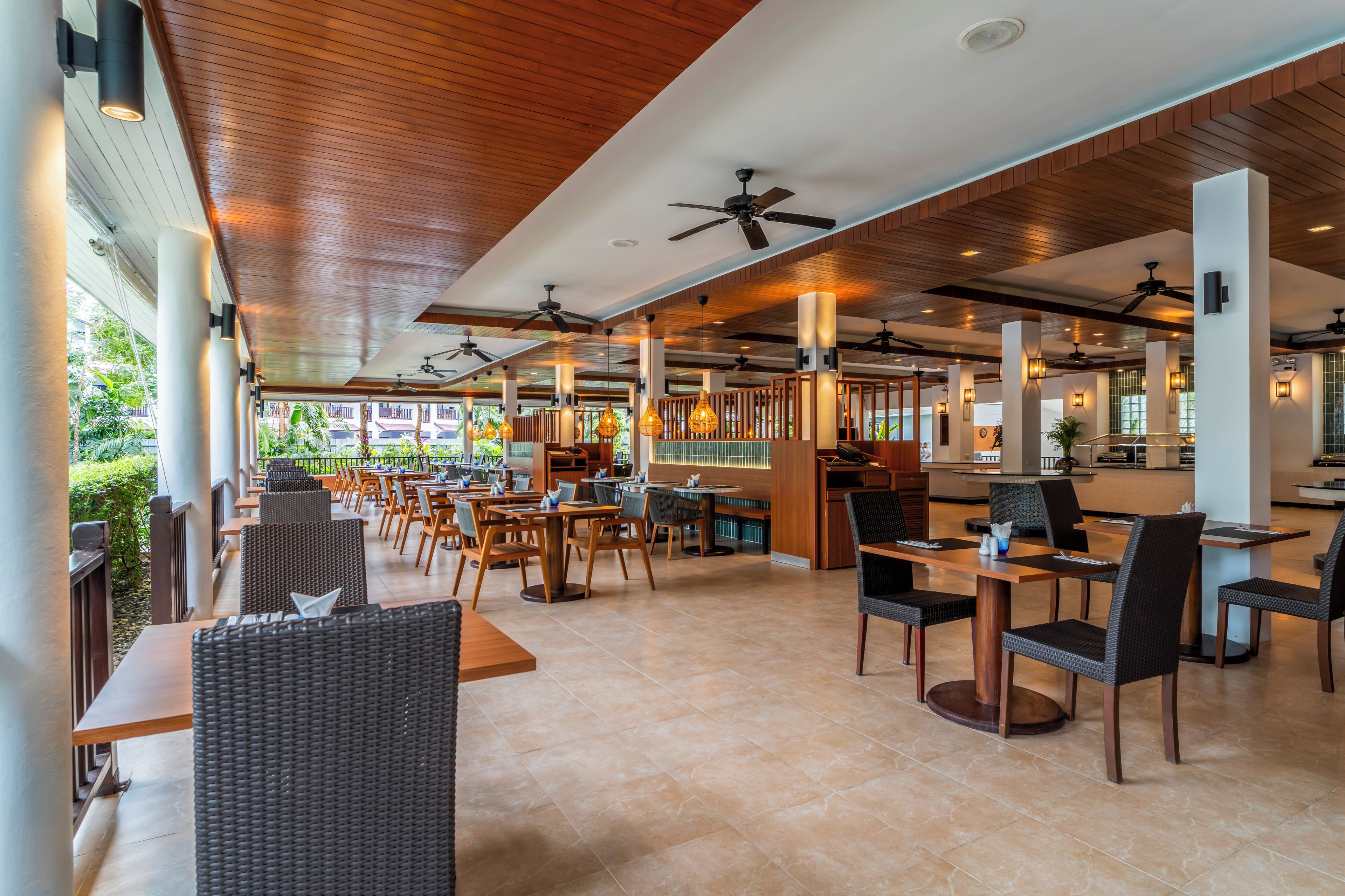 A spacious outdoor dining area with multiple tables and chairs, ceiling fans, and a view of greenery outside.