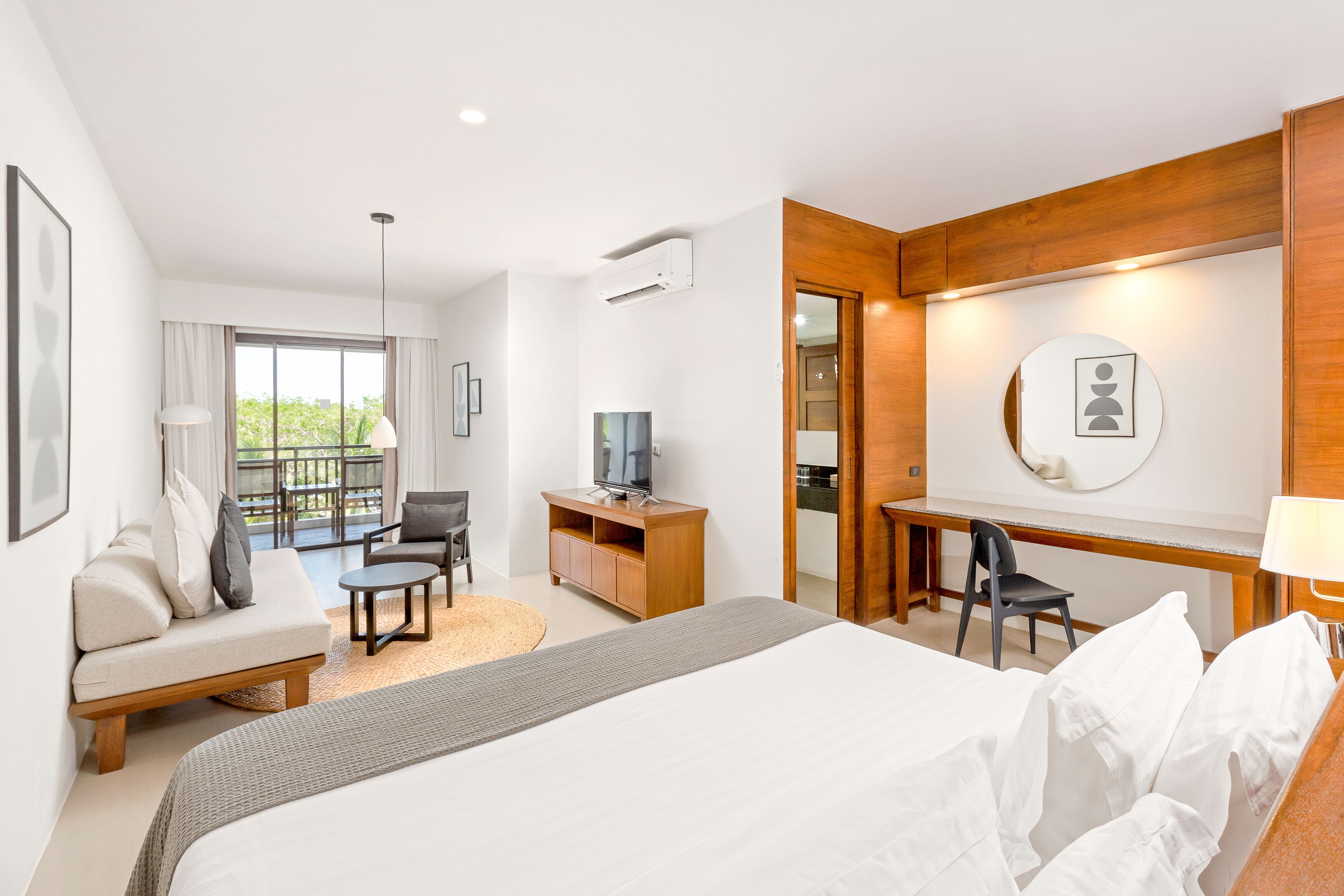A hotel room with a bed, a desk, a chair, a sofa, a TV, and a balcony with a view of greenery.