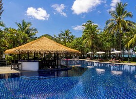 Ein Swimmingpool mit Bar unter einem Reetdach, umgeben von Palmen und Liegestühlen.