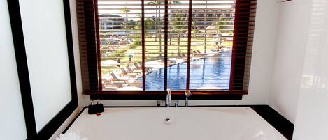 A bathroom with a large bathtub, a window with blinds, and a view of a pool and outdoor area.