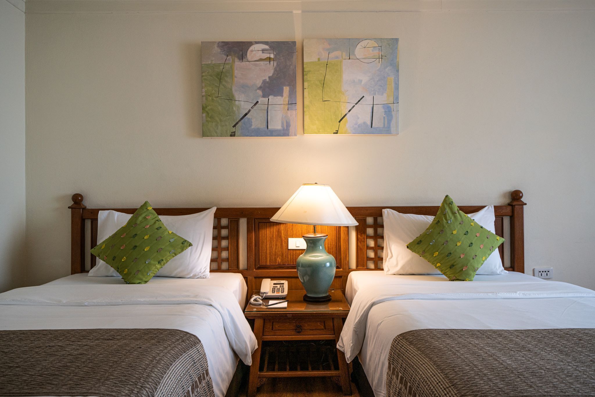 A hotel room with two beds, a nightstand with a lamp, a telephone, and two paintings on the wall.