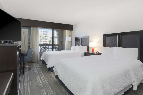 A hotel room with two beds, a desk, a chair, and a balcony with a view of buildings.