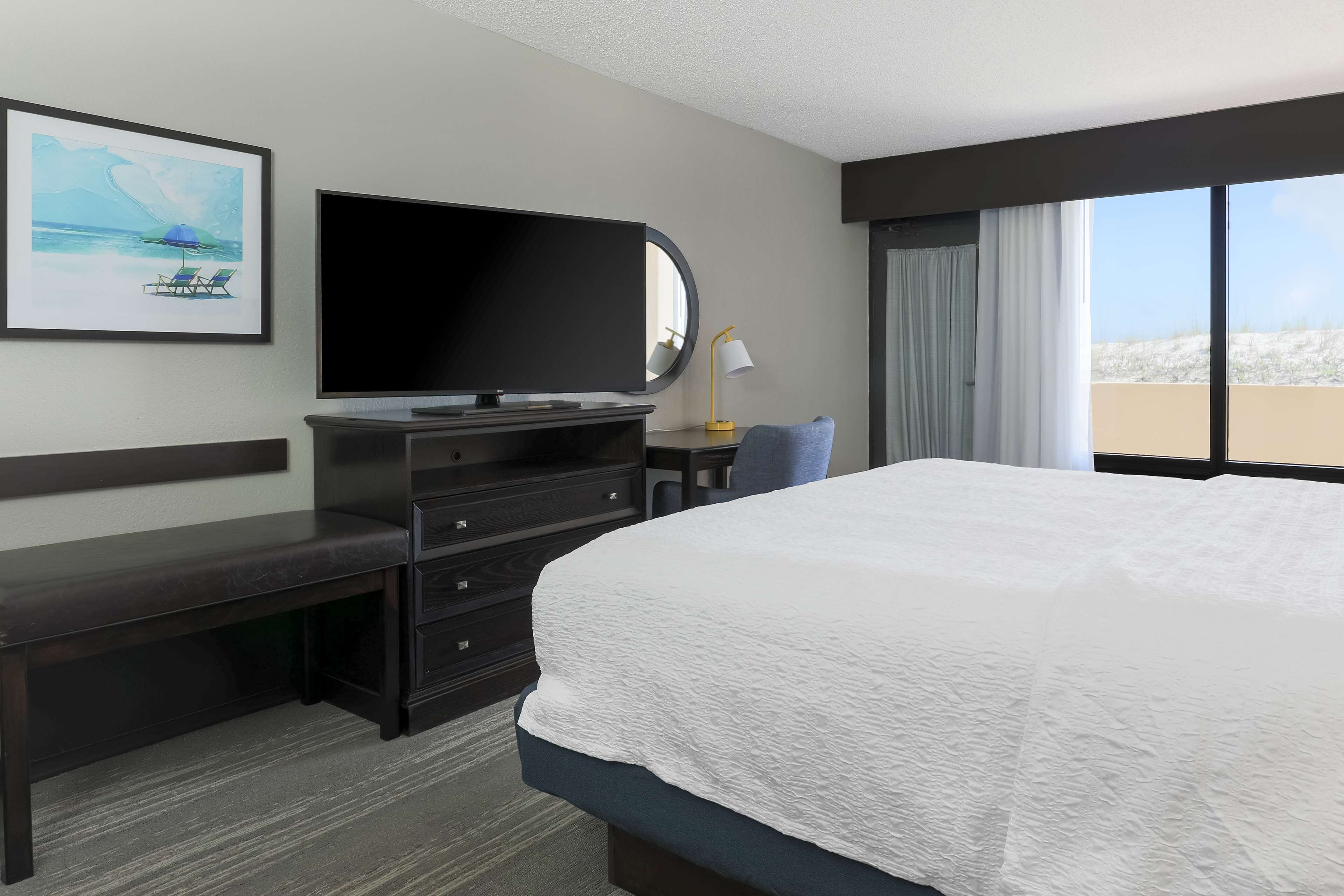 A hotel room with a large bed, a flat-screen TV, a desk with drawers, a chair, a desk lamp, and a large window with curtains.