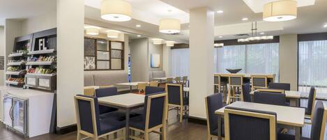A modern hotel dining area with tables and chairs, a bar area, and a view of the outdoors through large windows.
