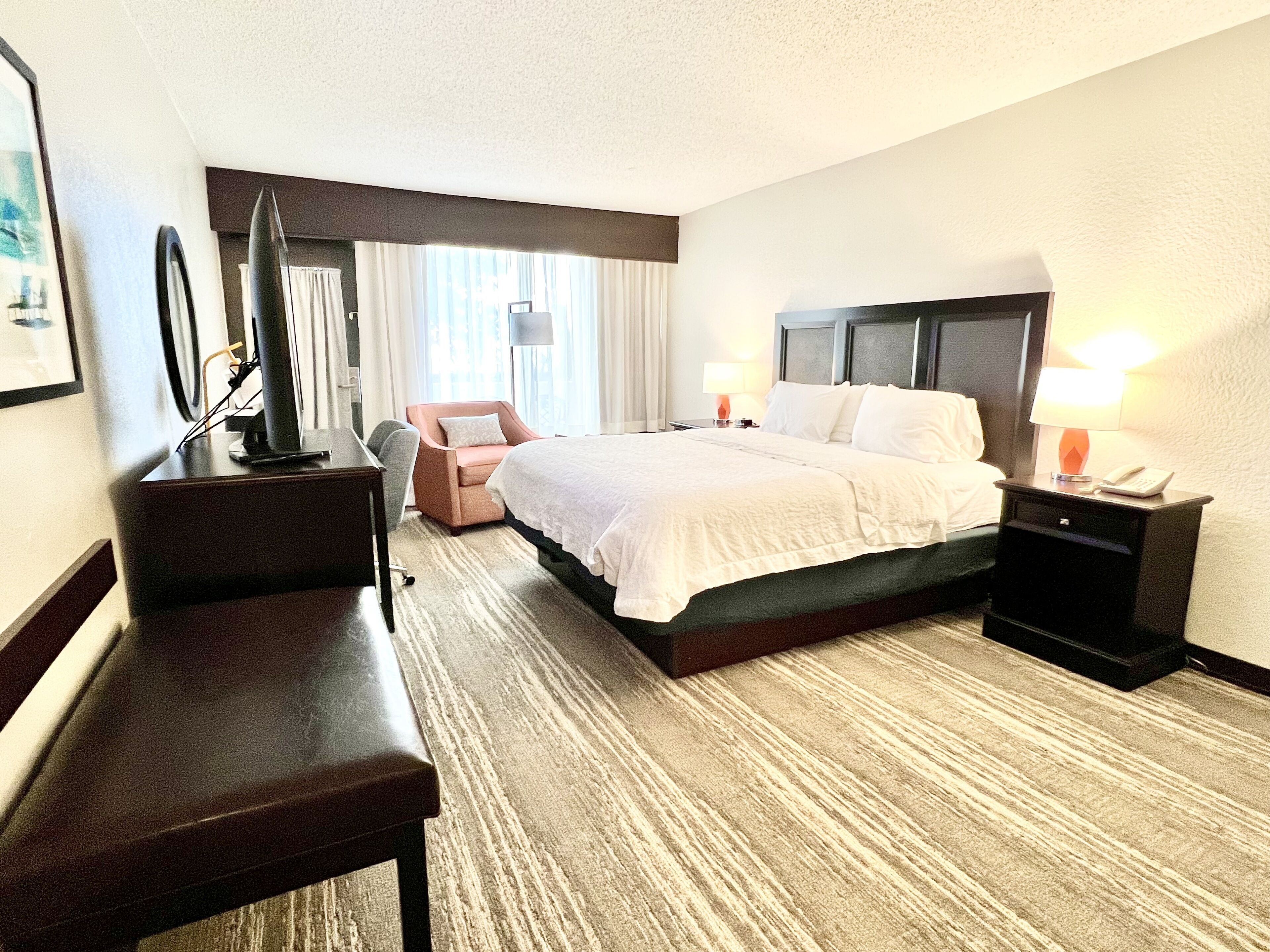 A hotel room with a bed, a desk, a chair, a TV, and a nightstand with a lamp and phone.