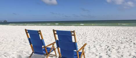 Two blue beach chairs on a sandy beach with a clear sky and ocean in the background.