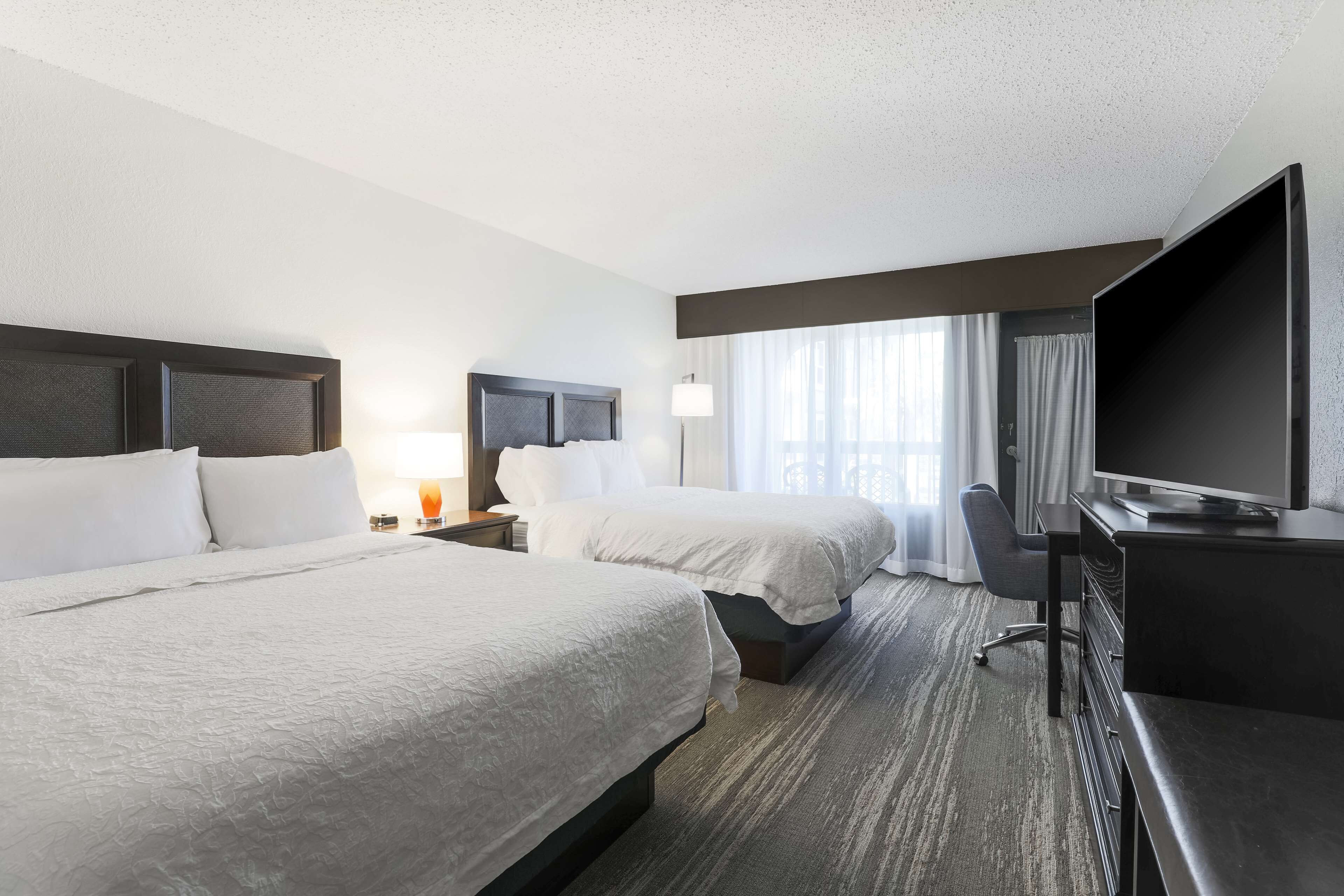 Quarto de hotel com duas camas, uma escrivaninha, televisão e janela com cortinas.