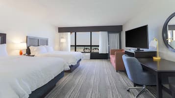 A hotel room with two beds, a desk with a chair, a television, and a large window with curtains.