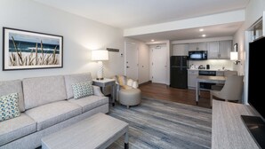 A modern living room with a sofa, ottoman, and coffee table. A kitchen with a microwave and refrigerator is visible in the background. - Hampton Inn and Suites Amelia Island Historic Harbor Front (Fernandina Beach)