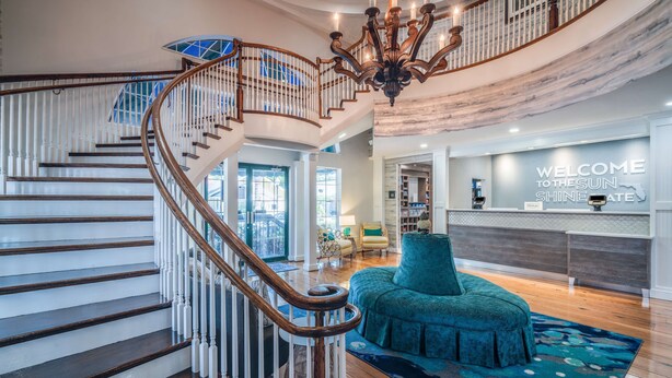 A grand staircase with a wooden handrail and white balusters, leading to a second floor. A chandelier hangs from the ceiling, and a sign reads "WELCOME TO THE SUN SHINE STATE". - Hampton Inn and Suites Amelia Island Historic Harbor Front (Fernandina Beach)