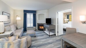 A modern hotel room with a sofa, armchair, and a dining table. A fireplace with a TV is visible in the background. - Hampton Inn and Suites Amelia Island Historic Harbor Front (Fernandina Beach)
