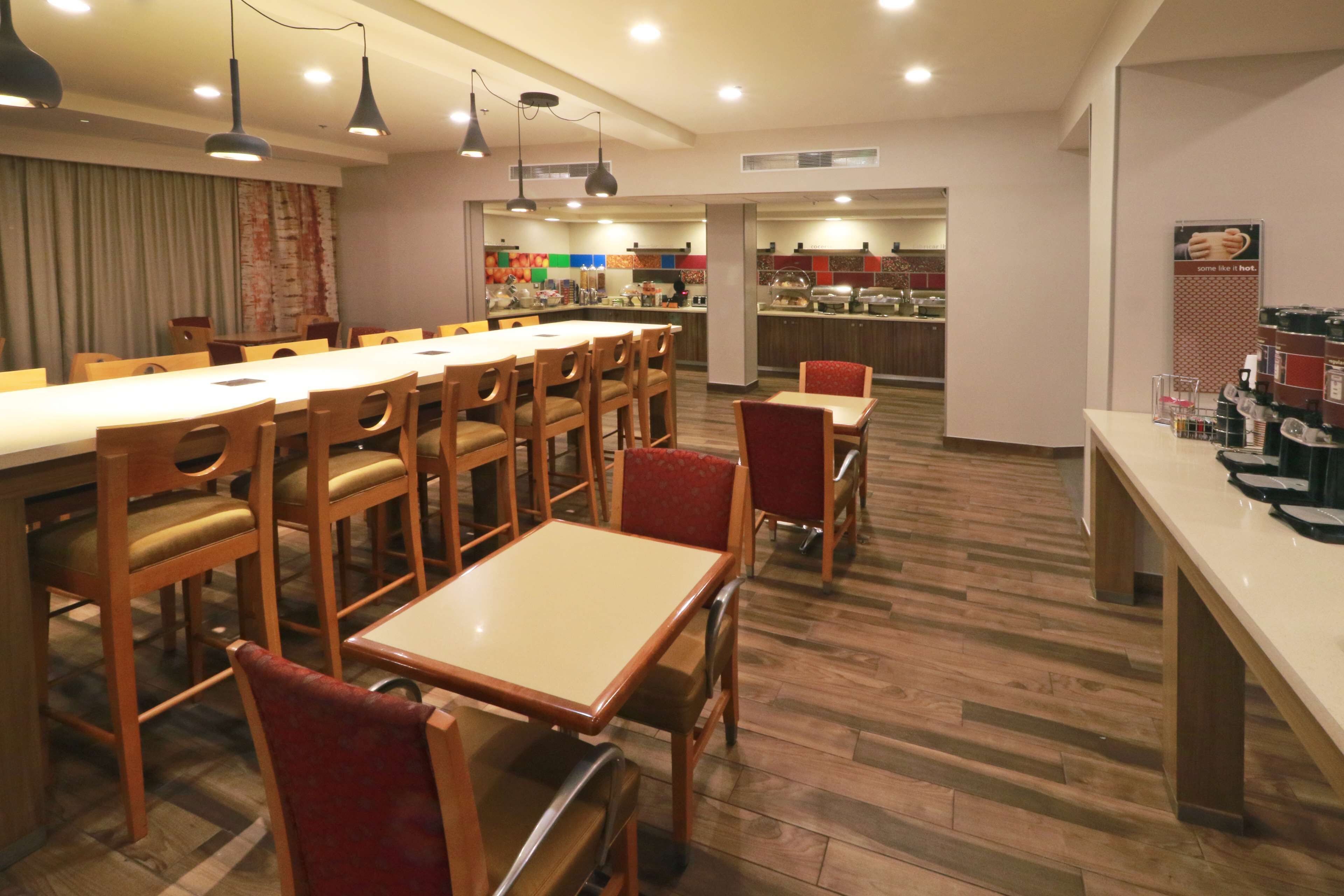 A hotel dining area with multiple tables and chairs, a kitchen in the background, and a counter with various items.