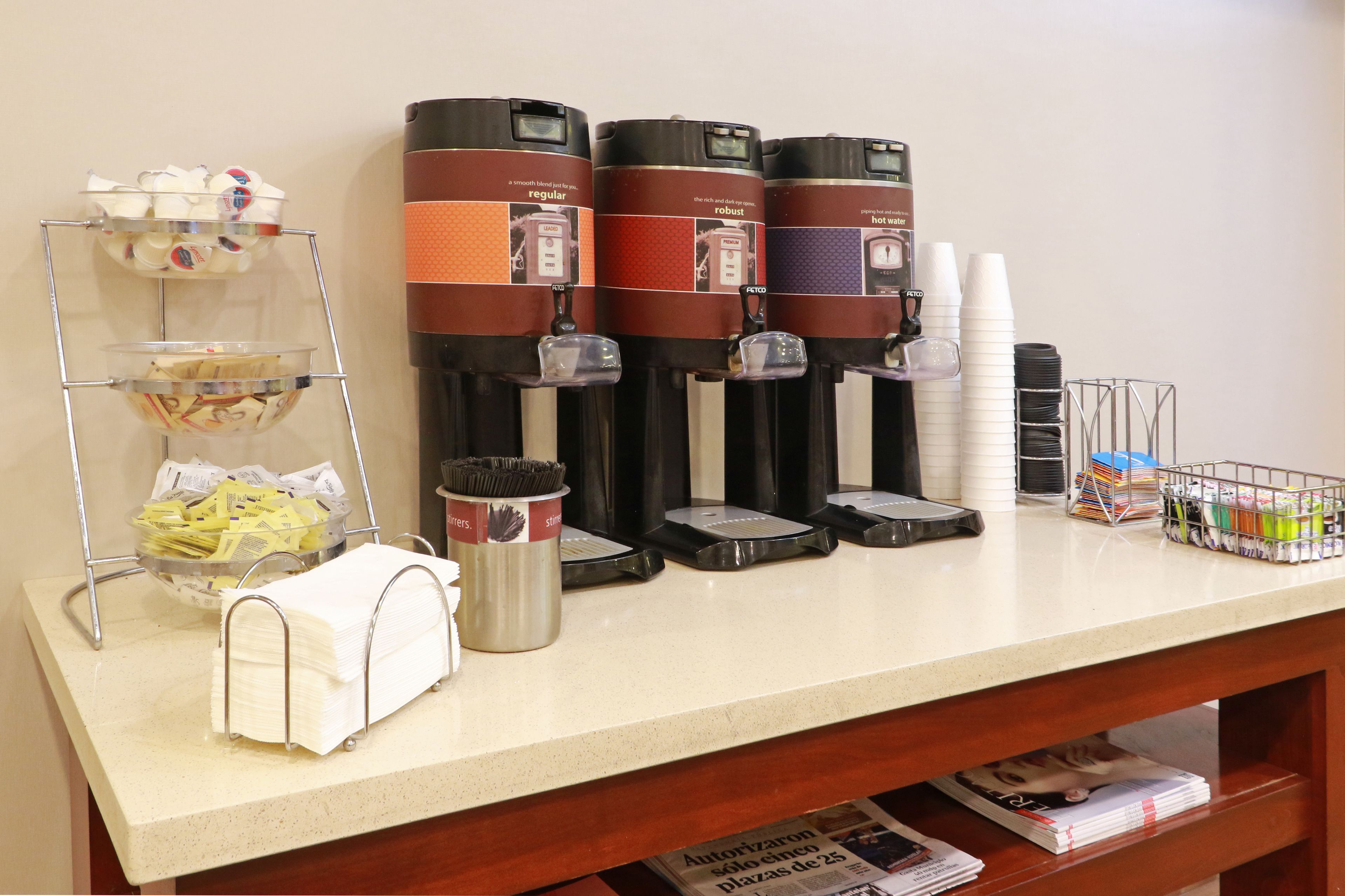 Una estación de autoservicio para bebidas con varios dispensadores de café, un dispensador de toallas de papel y un portarrollos de papel.