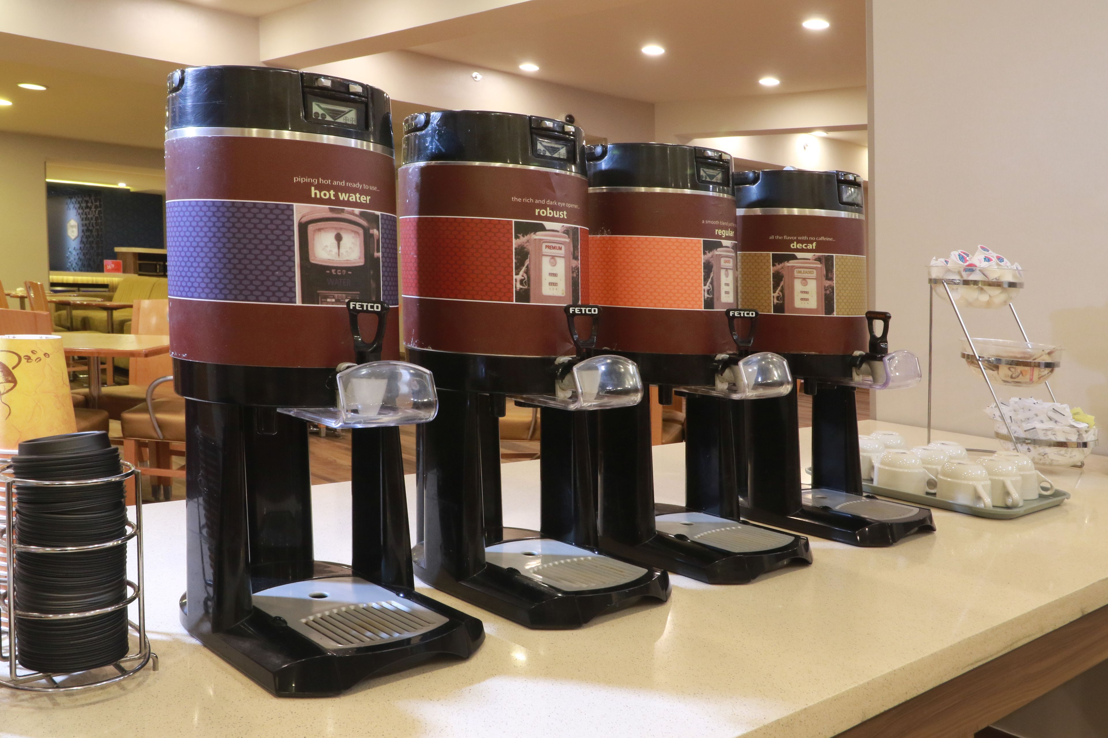 A row of coffee machines with hot water dispensers, a coffee station, and a tray of disposable cups.
