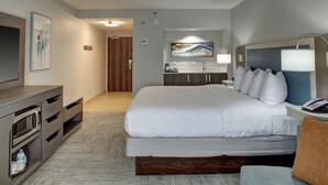 Habitación de hotel con cama, escritorio con lámpara, televisor, baño con lavamanos y espejo, y un cuadro en la pared.