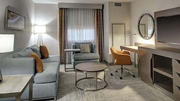 A modern hotel room with a grey sofa, a round coffee table, a desk with a chair, a TV, and a large window with curtains.