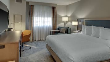 A hotel room with a large bed, a desk with a chair, a small table, a lamp, and a TV.