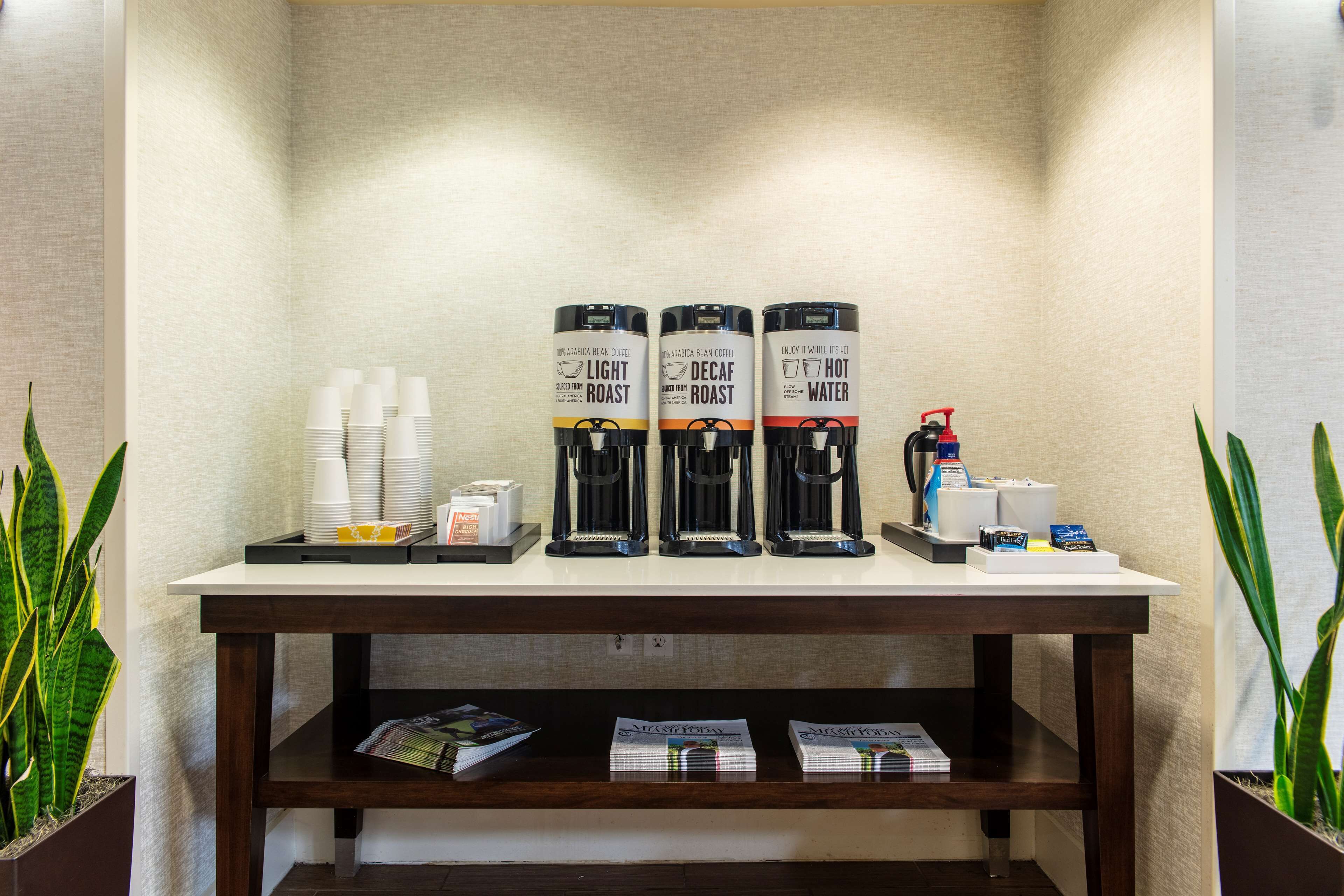 A coffee station with three coffee makers labeled "Light Roast," "Decaf Roast," and "Hot Water." There are stacks of white cups and a small tray with a coffee pot and a red fire extinguisher.