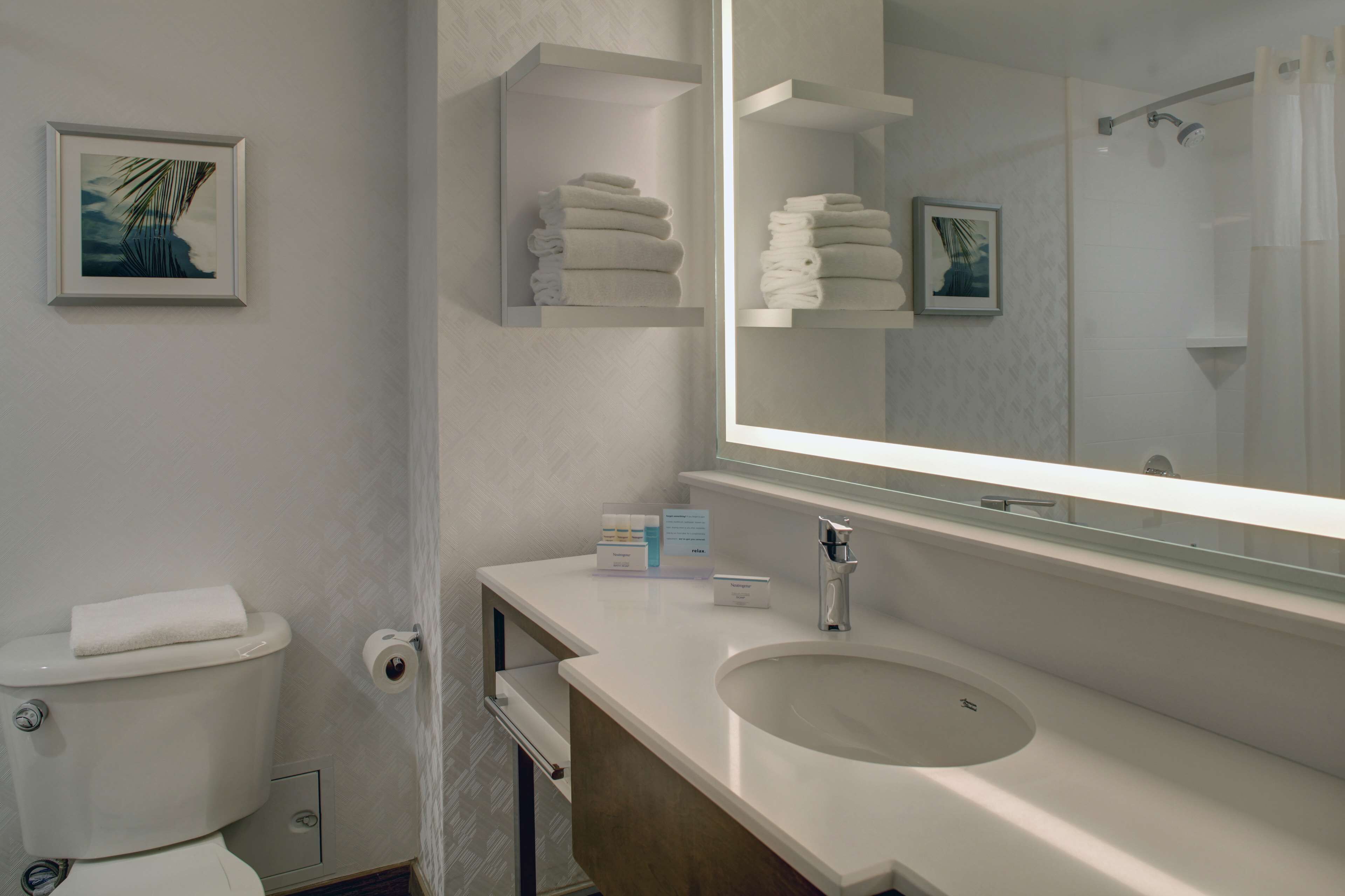 A modern bathroom with a toilet, sink, and mirror. There are towels on a shelf and a framed picture on the wall.
